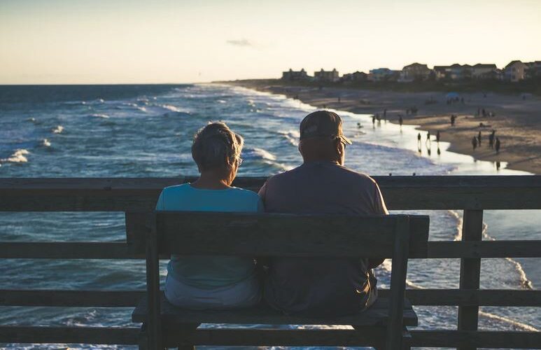 Old Couple Sitting On Bench At Beach